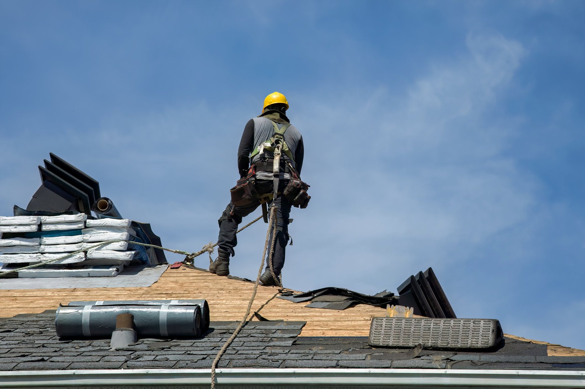 Construction worker using pneumatic nailer and safety helmet applying roofing materials against blue sky backdrop