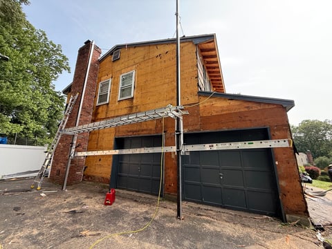 Red brick house under renovation with two black garage doors, metal scaffolding, and construction equipment visible