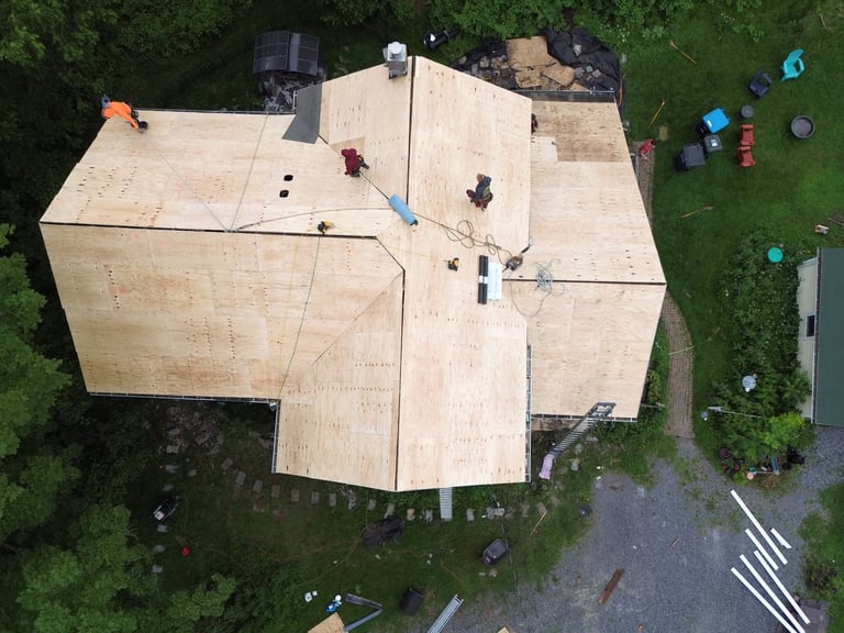 Aerial view of a large plywood house structure under construction with workers and equipment on a wooden roof, surrounded by green lawn