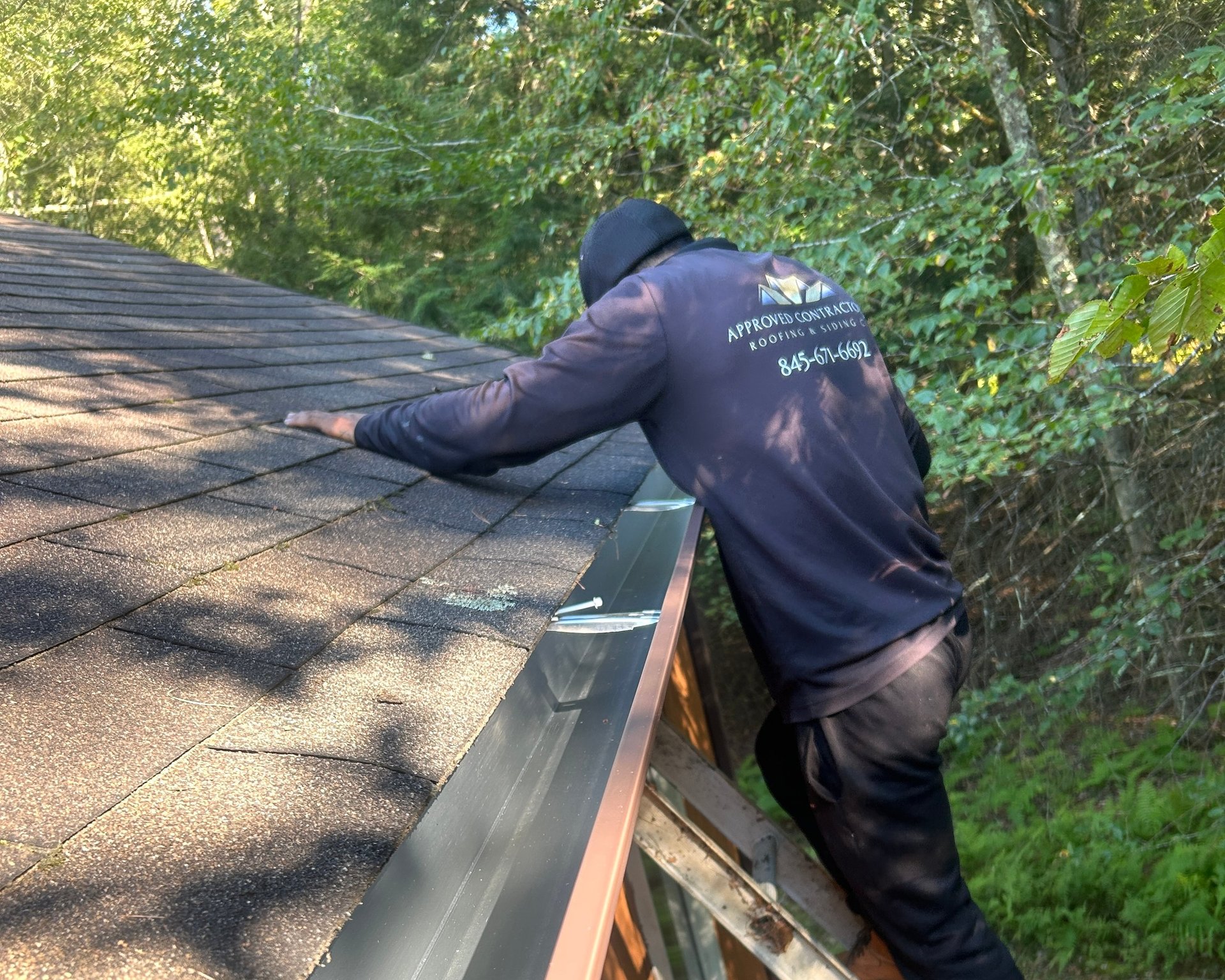 Worker in black jacket installing or inspecting roofing material on a residential roof surrounded by trees