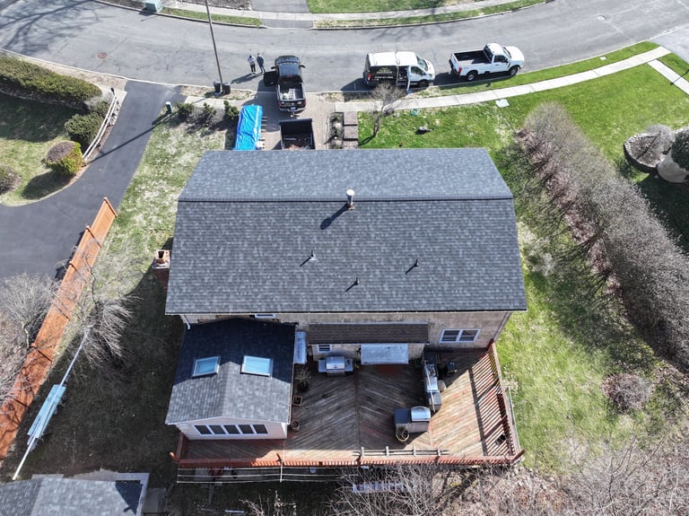 Aerial view of a residential house with gray roof, wooden deck, and driveway with parked vehicles surrounded by landscaping and grass