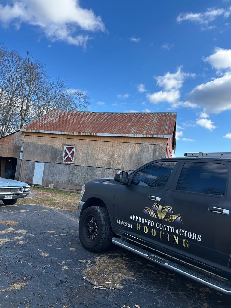 Black pickup truck labeled Approved Contractors Roofing parked in front of a weathered barn with red metal roof