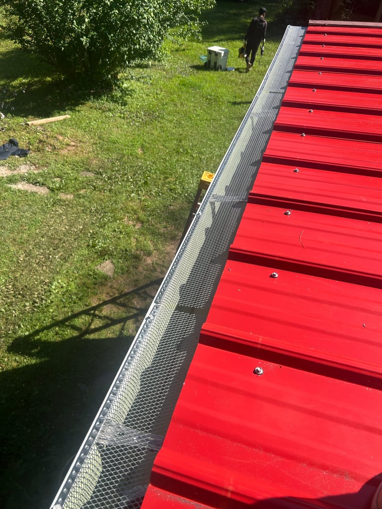 Aerial view of a red metal roof with gutter and mesh guard overlooking a green yard with people and trees