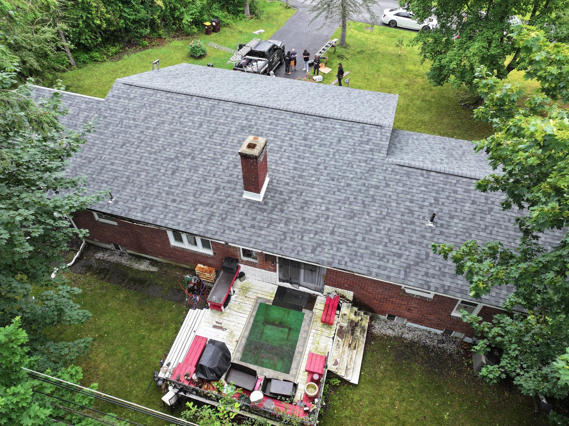 Aerial view of red brick house with gray roof surrounded by green trees and emergency vehicles in driveway