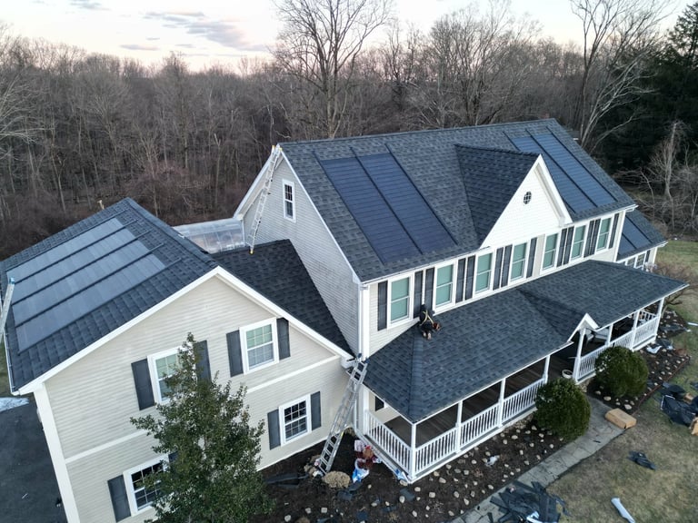 Aerial view of a modern two-story farmhouse with white siding, dark blue roof, and wraparound porch surrounded by bare trees