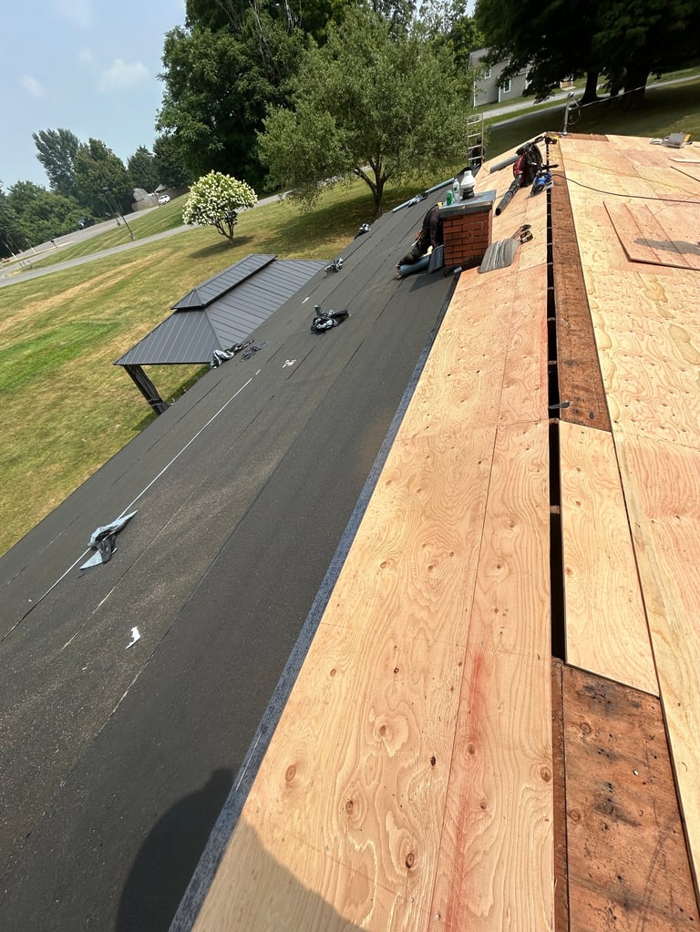 Aerial view of roof with workers installing dark shingles and plywood, residential house with green lawn and trees visible