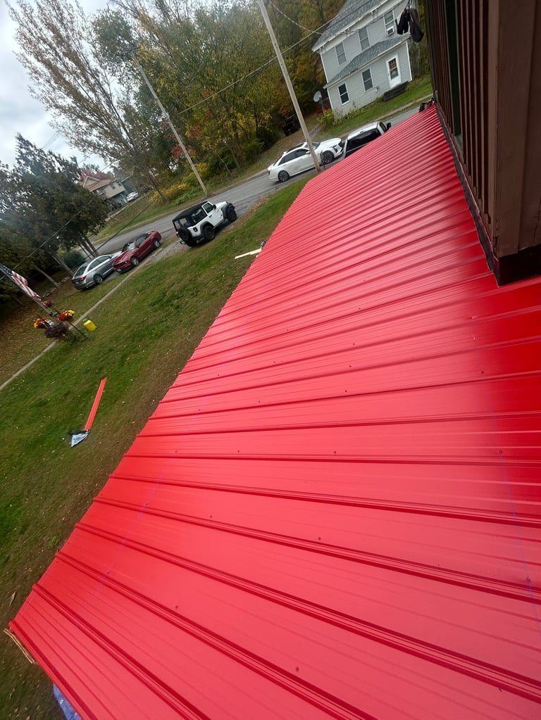 Elevated view of a bright red metal roof with horizontal ribbing overlooking a driveway, parked vehicles, and a white house surrounded by trees