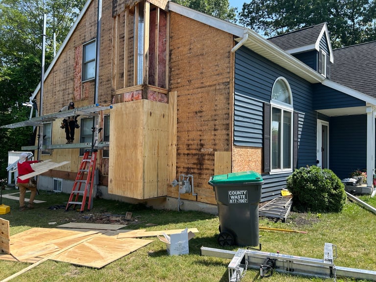 A tiny home under construction beside a blue house, with wooden framing, tools, and debris visible in the yard