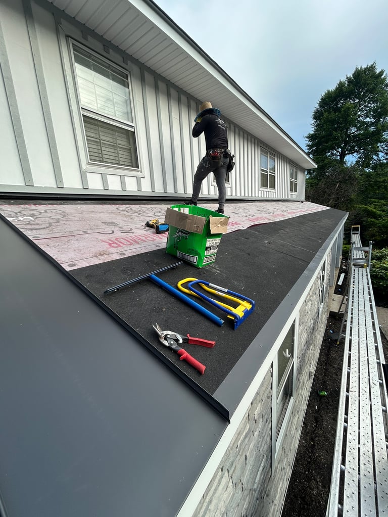 Worker in black clothing standing on a roof with tools including a green toolbox, hammers, and pliers laid out on black roofing material next to a white house