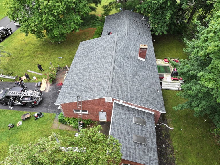 Aerial view of a brick house with gray shingled roof surrounded by green trees and lawn, with a driveway and patio visible