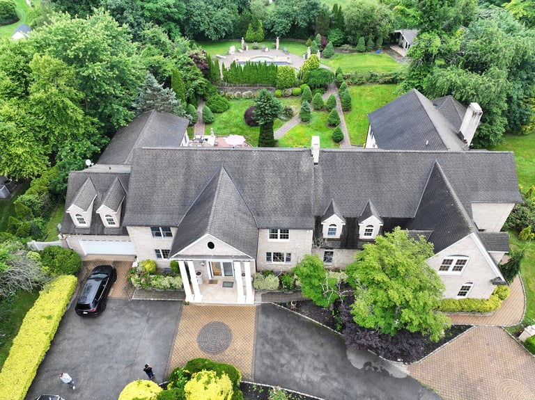 Aerial view of a large residential home with gray slate roof, manicured gardens, driveway, and surrounding trees