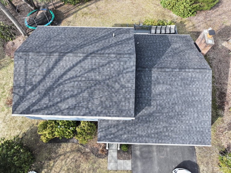Aerial view of a house with gray shingled roof, showing two peaked sections and surrounding landscaping
