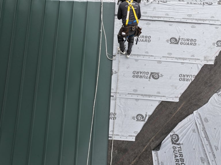 Worker in safety harness rappelling down green metal roof during construction installation
