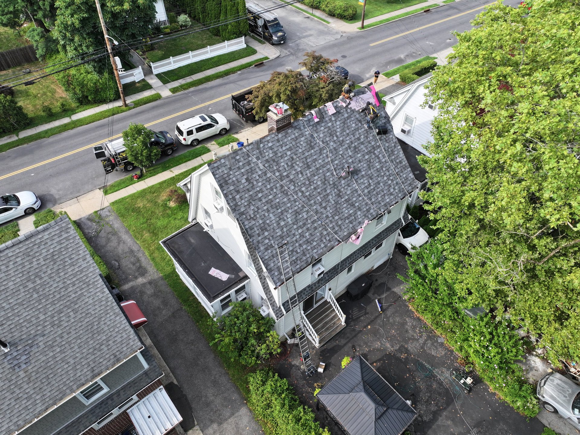 Aerial view of a white house with gray roof surrounded by trees and parked cars on a residential street